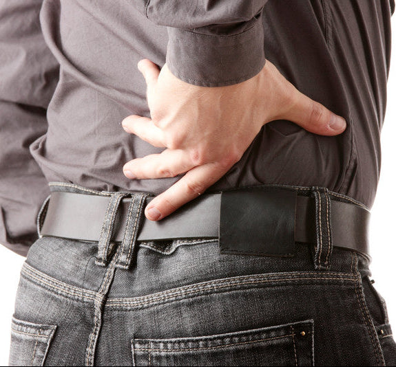 A close-up of a person from the back wearing a dark gray button-up shirt, black belt, and dark gray jeans, holding their lower back in pain against a white background.